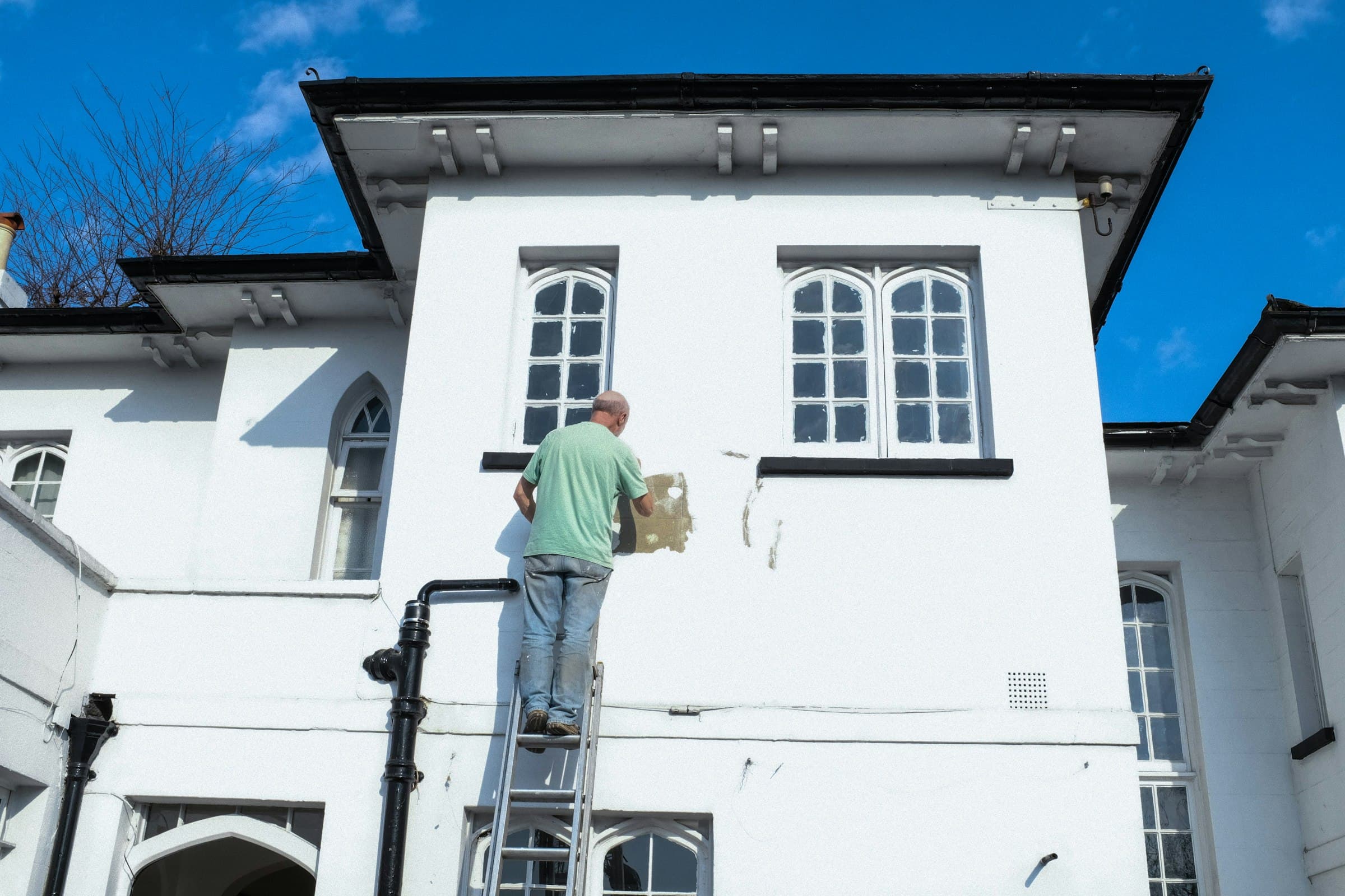 A painter on a ladder applying fresh paint to the exterior of a premium stucco home under a clear blue sky