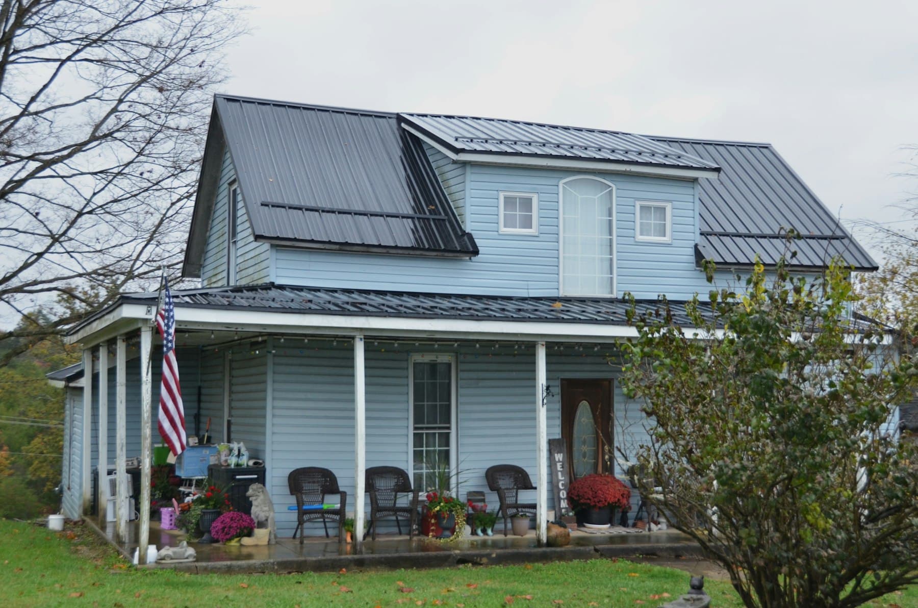 A light blue American farmhouse with a wraparound porch and an American flag
